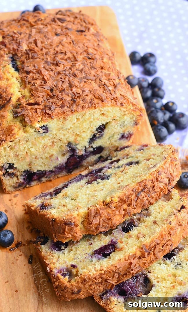 view from above of loaf of toasted coconut blueberry bread with the first 3 slices stacked in front of the rest of the loaf on a wooden cutting board with blueberries scattered around on table.