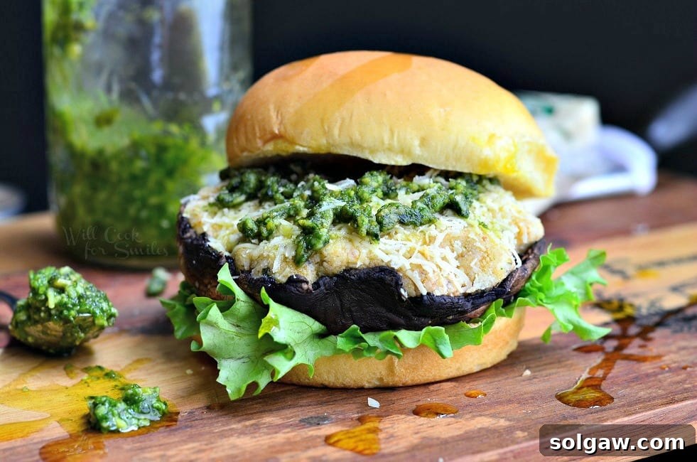 front view of Italian stuffed portobello burger on a bun and bed of lettuce sitting on a wooden board with a spoon of pesto at the bottom left and block of cheese in the background along with a bottle of pesto