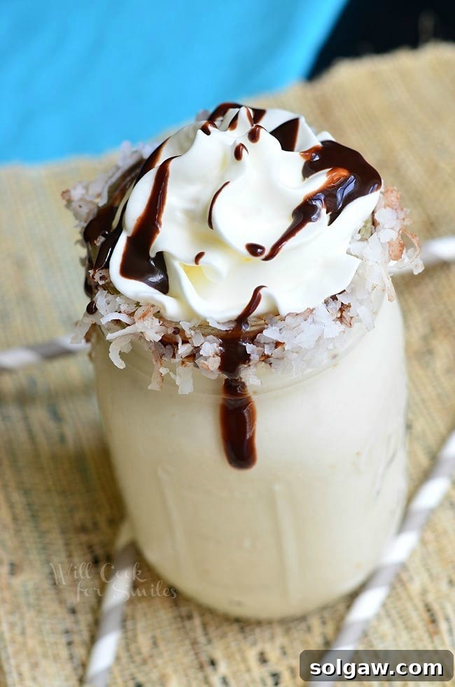 view from above of mason jar filled with lightened almond joy milkshake on a tan placemat with 3 straw around the base of the jar and a blue cloth in the background topped with whipped cream and chocolate syrup