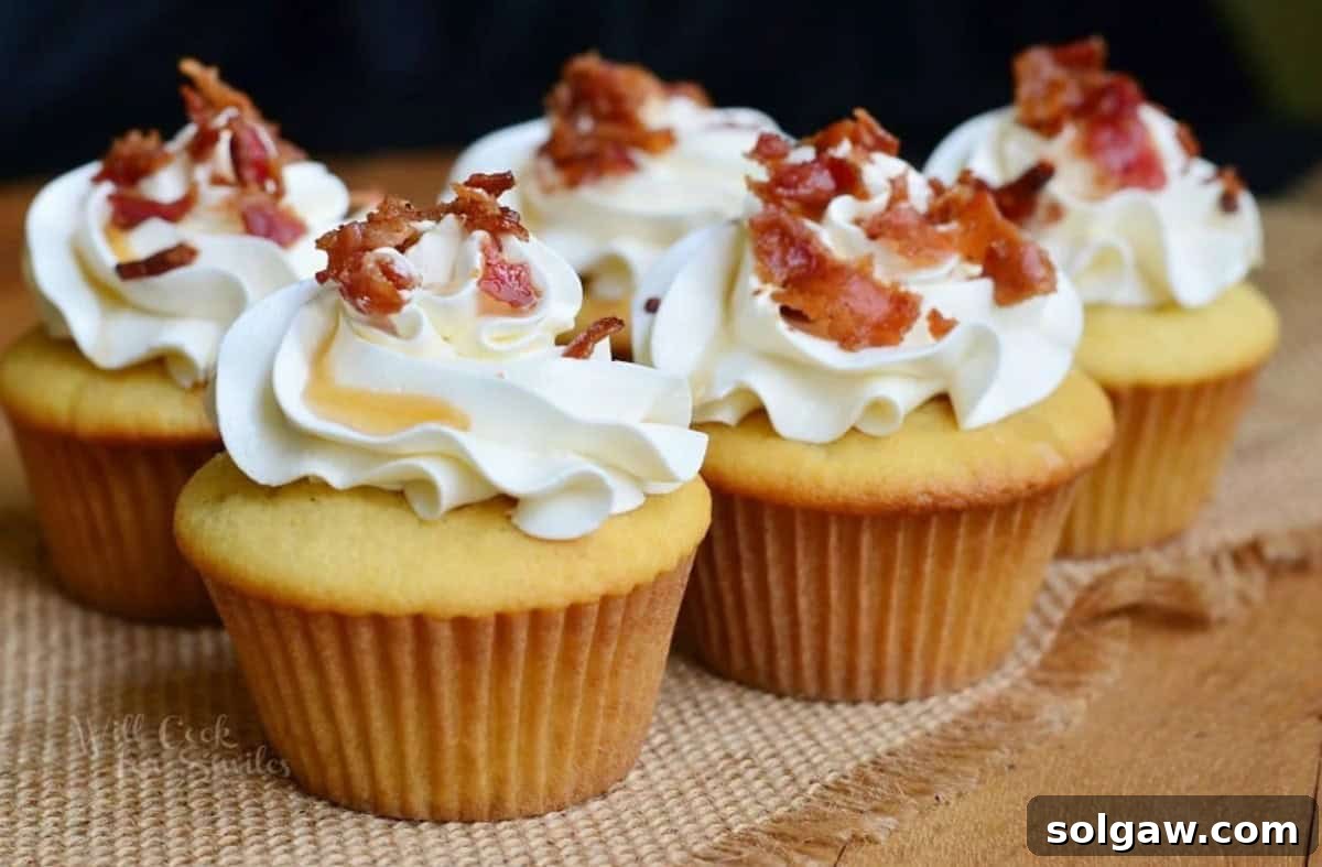 A row of several maple bacon cupcakes, each topped with a generous swirl of maple buttercream frosting, sprinkled with crispy bacon bits, and drizzled with maple syrup.