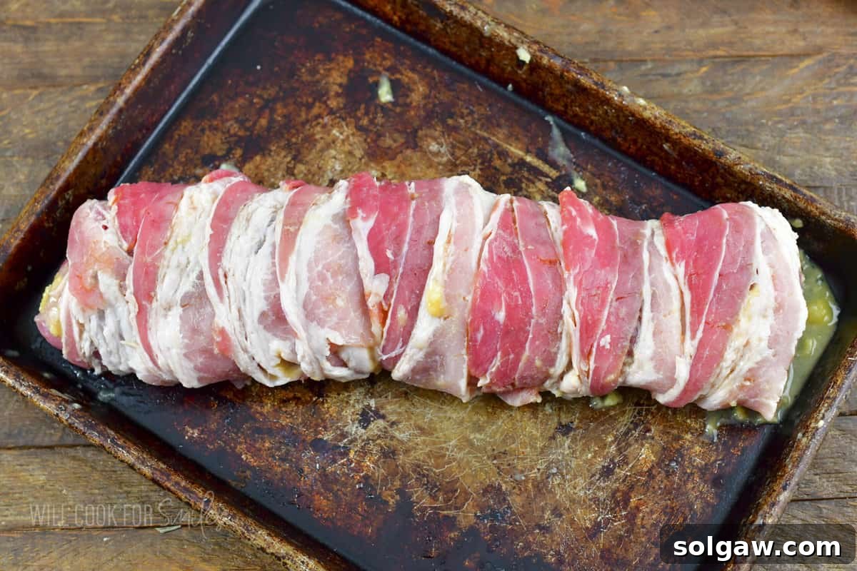 Bacon Wrapped Pork Perfection 6 Uncooked pork tenderloin meticulously wrapped in bacon and placed on a baking sheet.