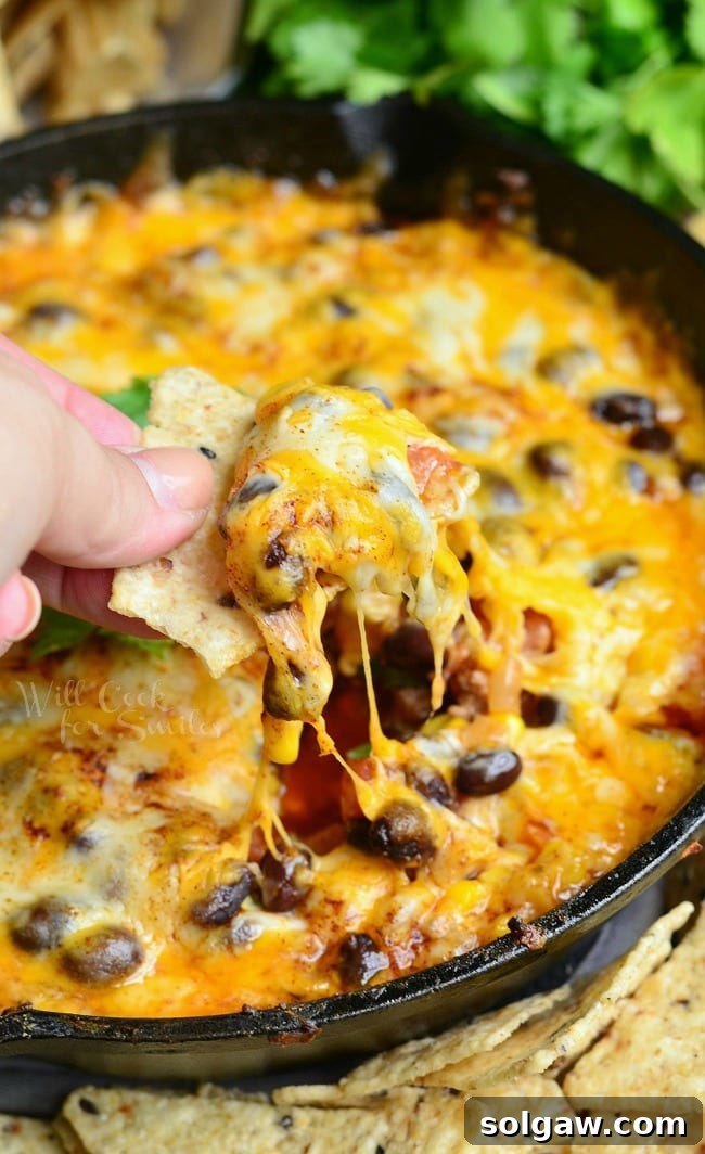 A black skillet with hot 7 layer dip rests on a table surrounded by chips and a hand dipping one chip into the dip