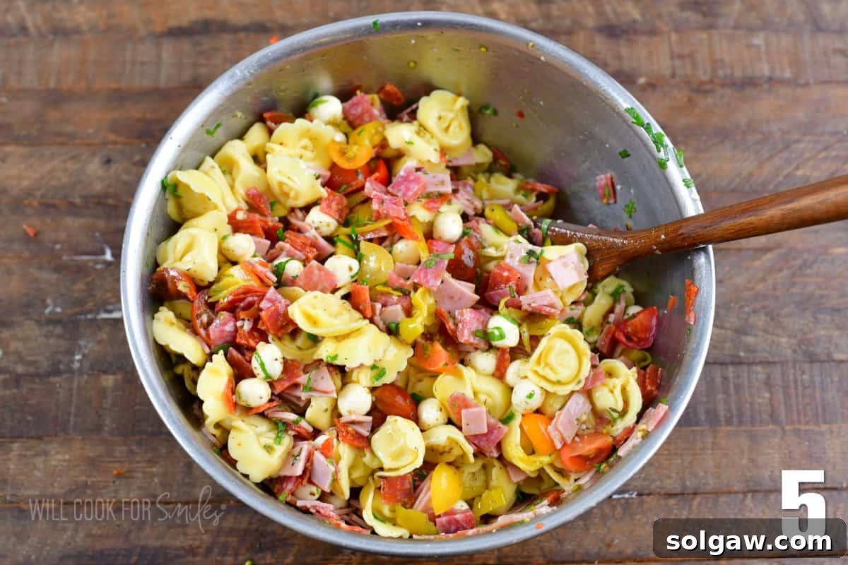 mixed Italian tortellini salad in a metal mixing bowl with a wooden spoon. A large bowl of freshly mixed tortellini salad.