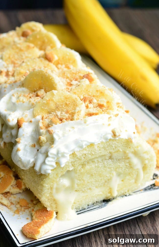 A close-up view of a Banana Pudding Cake Roll on a pristine white plate, garnished with whipped cream, crumbled vanilla wafers, and fresh banana slices. An unpeeled banana is visible in the background.