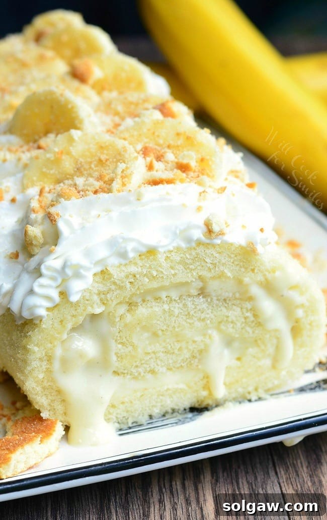 A beautifully presented Banana Pudding Cake Roll on a serving plate, topped with whipped cream, crushed vanilla wafers, and banana slices. An unpeeled banana is positioned to the right.