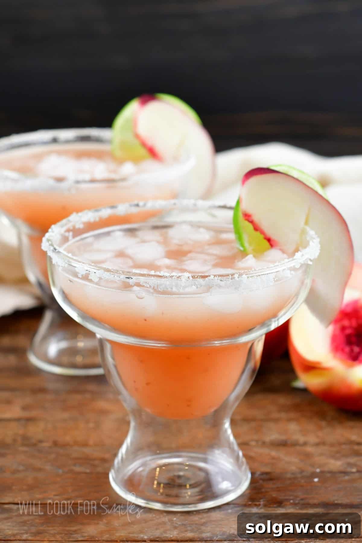 A vibrant peach margarita, garnished with fresh peach slices and a salted rim, sits on a wooden surface, promising refreshing delight.