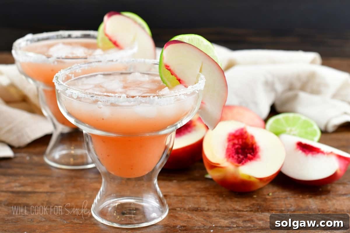 Two perfectly garnished peach margaritas in elegant glasses are placed alongside fresh peach slices on a light surface.